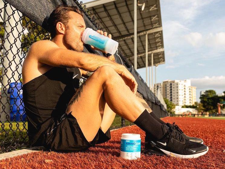 A man drinks AminoLean Energy blue raspberry pre workout powder. On right store shelf with other brands of supplements. 